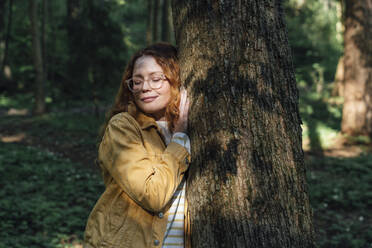 Smiling woman with eyes closed embracing tree in forest - VPIF06526