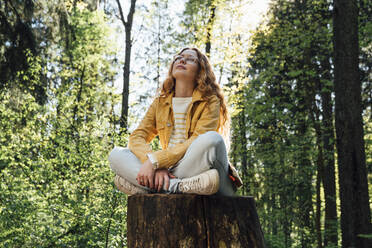 Thoughtful woman sitting cross-legged on tree stump in forest - VPIF06529