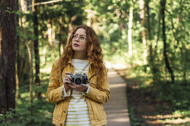 Contemplative woman holding camera in forest - VPIF06546