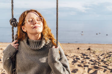 Frau mit geschlossenen Augen genießt das Sonnenlicht auf einer Schaukel am Strand - VPIF06560