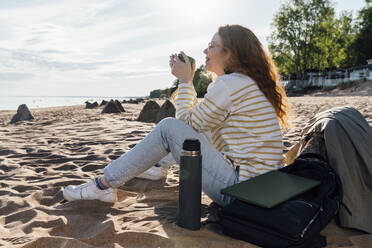 Lächelnde Frau trinkt Kaffee in einem isolierten Becher am Strand - VPIF06577