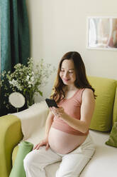 Happy pregnant woman using smart phone on sofa at home - SEAF00971