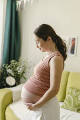 Schwangere Frau mit Unterleib am Sofa stehend zu Hause - SEAF00973