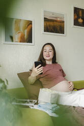 Lächelnde schwangere Frau, die zu Hause ein Mobiltelefon benutzt - SEAF00986