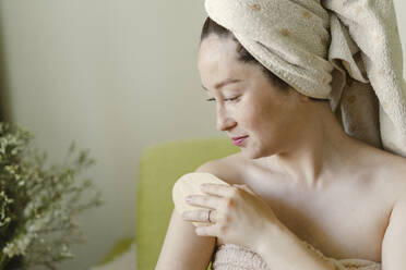 Smiling woman massaging with dry brush sitting on sofa at home - SEAF00992