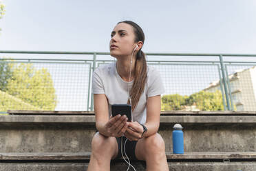 Nachdenkliche Frau mit Handy in der Hand hört Musik über In-Ear-Kopfhörer - FMOF01512