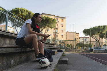 Frau mit Fußball hört Musik über In-Ear-Kopfhörer auf einer Bank am Sportplatz - FMOF01514