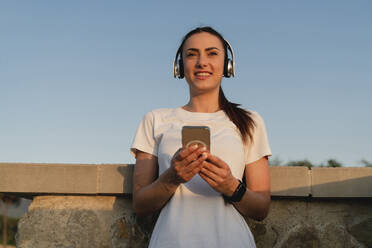 Smiling woman holding mobile phone listening music through wireless headphones in front of sky - FMOF01543