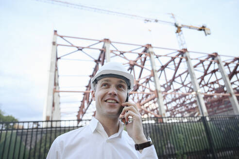 Glücklicher Ingenieur, der mit seinem Smartphone im Baugewerbe telefoniert - EYAF01933