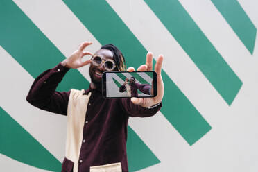 Lächelnder Mann zeigt Selfie auf Smartphone-Bildschirm vor gestreifter Wand - ASGF02440