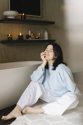 Lächelnde Frau sitzt auf dem Boden neben einer Badewanne im Badezimmer - SEAF01018