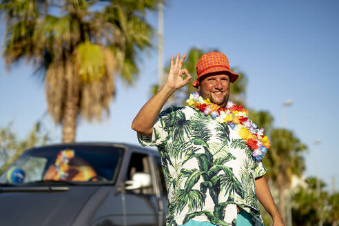Smiling man wearing hat and floral garland gesturing OK sign on sunny day - OCMF02458