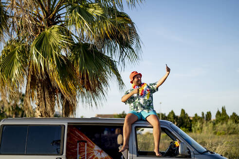 Happy man taking selfie through mobile phone sitting on top of van - OCMF02465