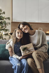 Glückliche Frau mit Weihnachtsgeschenk umarmt Tochter im Wohnzimmer - ABIF01707