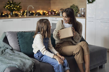 Lächelnde Frau zeigt ihrer Tochter, die zu Hause auf dem Sofa sitzt, ein Geschenk - ABIF01712