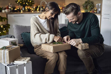 Lächelnde Frau mit Mann, der ein Weihnachtsgeschenk verpackt, sitzt zu Hause auf dem Sofa - ABIF01717