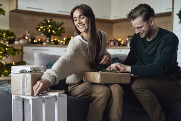Mann wickelt Geschenkbox von Frau im Wohnzimmer zu Weihnachten - ABIF01718