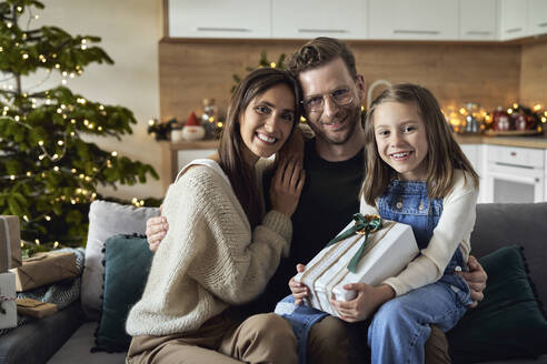 Smiling girl holding Christmas gift sitting with parents on sofa at home - ABIF01721