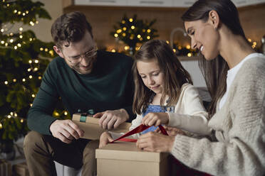 Girl opening Christmas present sitting with parents at home - ABIF01727