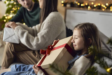Smiling girl embracing gift box sitting by parents at home - ABIF01732