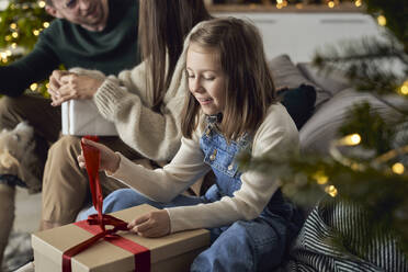 Lächelndes Mädchen öffnet Weihnachtsgeschenk der Eltern im Wohnzimmer - ABIF01733