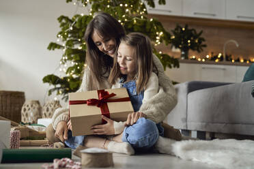 Girl holding Christmas present sitting with mother on floor at home - ABIF01739