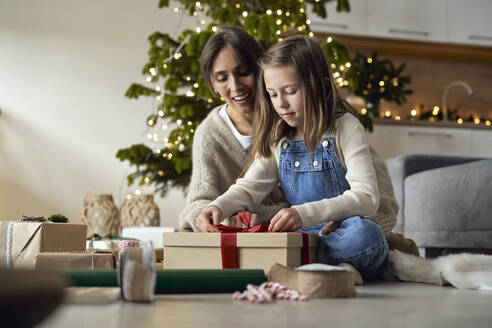 Mädchen, das mit seiner Mutter zu Hause auf dem Boden sitzend ein Geschenk verpackt - ABIF01741