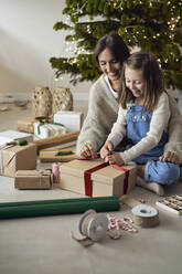 Smiling woman with daughter packing gift sitting on floor at home - ABIF01742