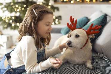 Girl looking at dog wearing antler sitting on sofa at home - ABIF01751