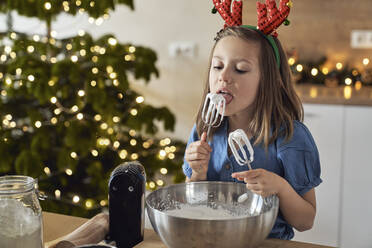 Girl licking cookie dough from whisk standing in kitchen - ABIF01756