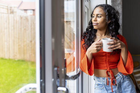 Nachdenkliche Frau steht mit einer Tasse Kaffee am Fenster - WPEF06020