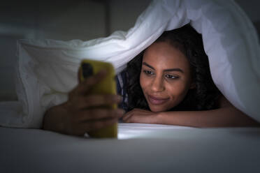 Junge Frau zugedeckt unter einer Decke, die im Schlafzimmer ihr Smartphone benutzt - WPEF06031