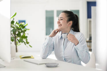 Happy businesswoman having coffee at desk in office - JOSEF10537