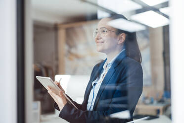 Lächelnde Geschäftsfrau mit Tablet-PC schaut durch ein Fenster - JOSEF10561