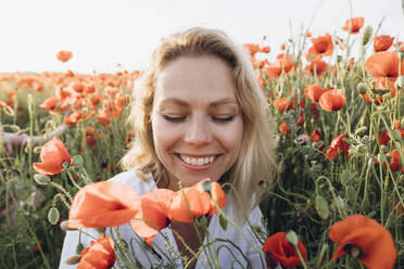 Glückliche Frau mit geschlossenen Augen sitzt inmitten roter Blumen auf einem Feld - SIF00188