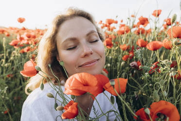 Smiling woman with eyes closed sitting amidst red flowers in field - SIF00189