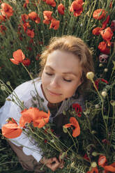 Frau mit geschlossenen Augen sitzt inmitten roter Blumen auf einem Feld - SIF00190