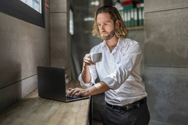 Serious businessman having coffee using laptop in office - DLTSF02942
