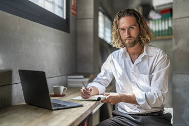 Seriöser Geschäftsmann mit Stift und Tagebuch am Laptop am Tisch im Büro - DLTSF02945