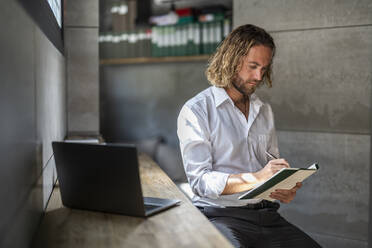 Ein leitender Angestellter schreibt in sein Tagebuch, während er an einem Tisch im Büro am Laptop sitzt - DLTSF02947