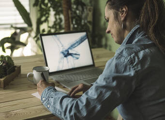 Freelancer making notes with wind turbine sketch on laptop screen at home office - UUF26493