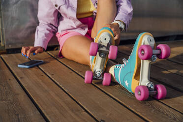 Woman wearing roller skates using mobile phone sitting on floorboard - VPIF06599