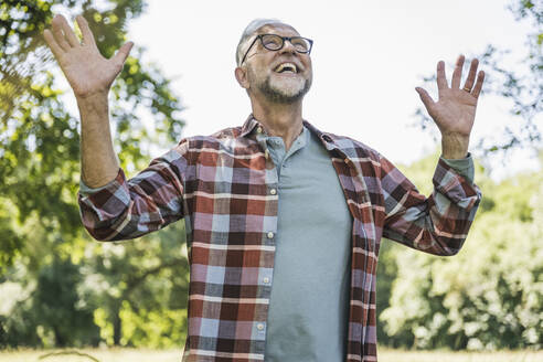 Cheerful senior man with arms raised standing in park - UUF26526