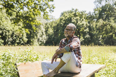 Smiling senior man wearing eyeglasses sitting on picnic table - UUF26532