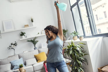 Glückliche Frau mit erhobenem Arm, die eine Sprühflasche hält und sich an der Reinigung ihres Hauses erfreut - JSRF02100