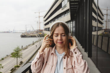 Lächelnde junge Frau mit drahtlosen Kopfhörern auf einem Balkon - IHF00920