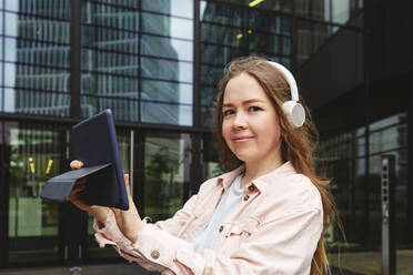 Happy woman wearing wireless headphones standing with tablet PC in front of building - IHF00926