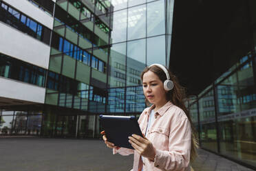 Young woman wearing wireless headphones using tablet PC in front of glass building - IHF00935