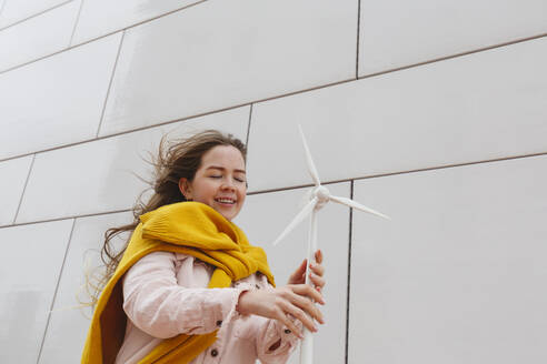 Lächelnde junge Frau mit Windradmodell vor einer Wand stehend - IHF00940