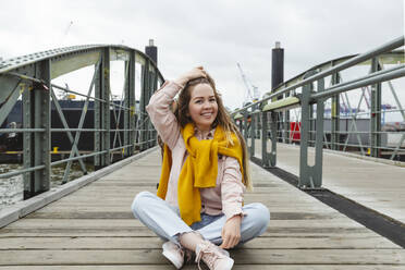 Happy young woman with hand in hair sitting on footbridge - IHF00948